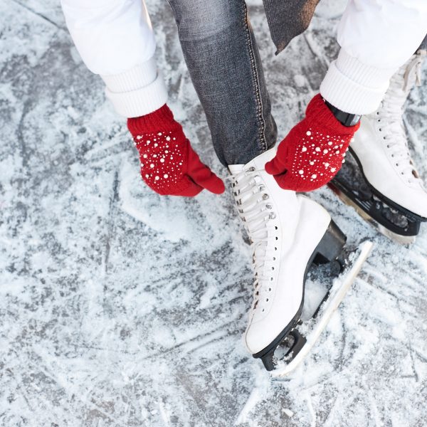 A close up shot of a person wearing red gloves lacing up a pair of white ice skates while sitting on ice