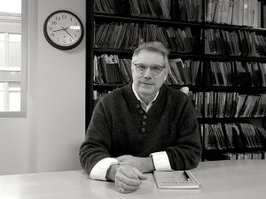 Middle-aged man with glasses and sleeves rolled up sitting at a table with a notebook and pen on it.