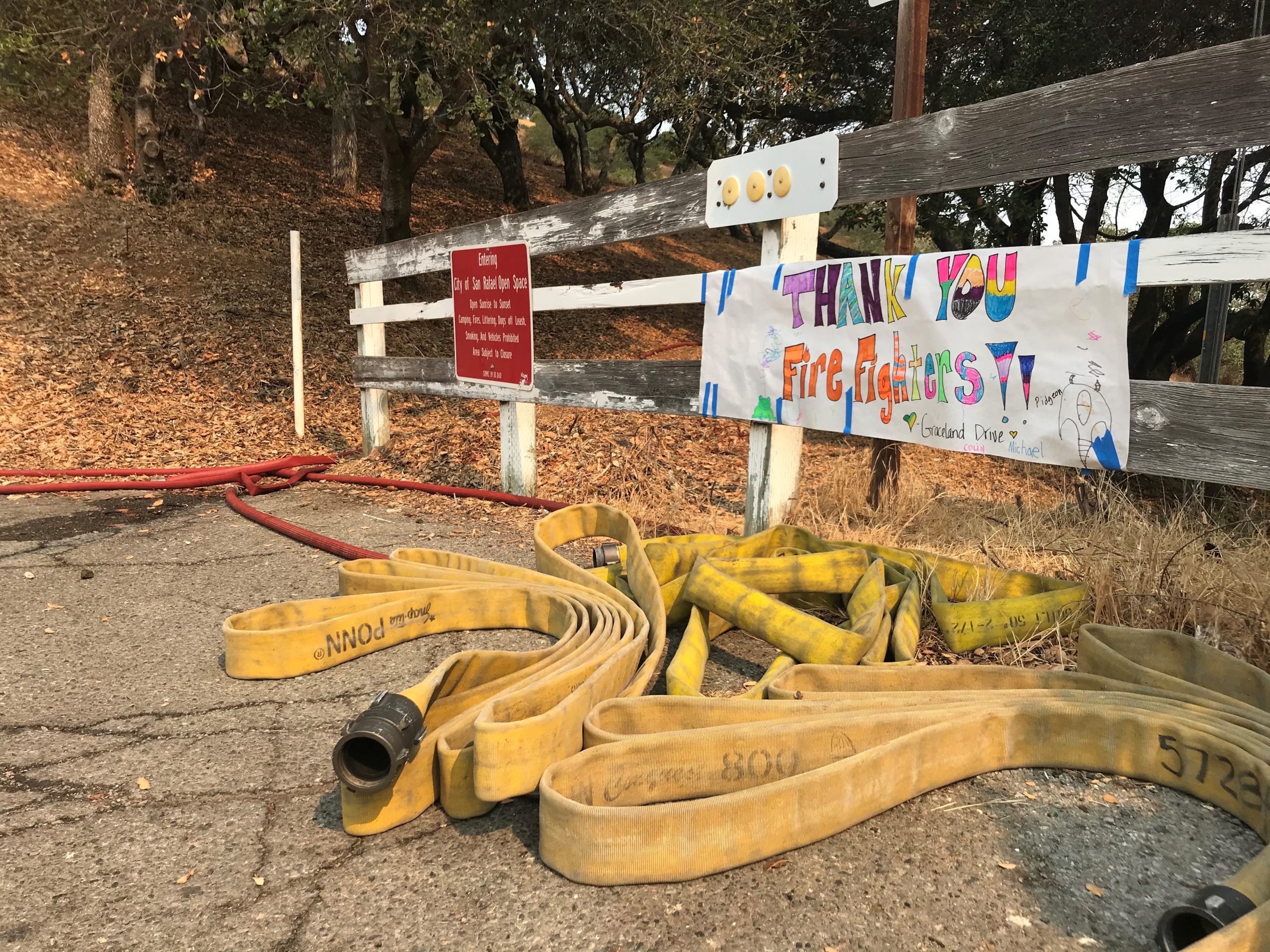 fire hose lays on the group in front of a hand written sign on a white fence that says Thank You Firefighters