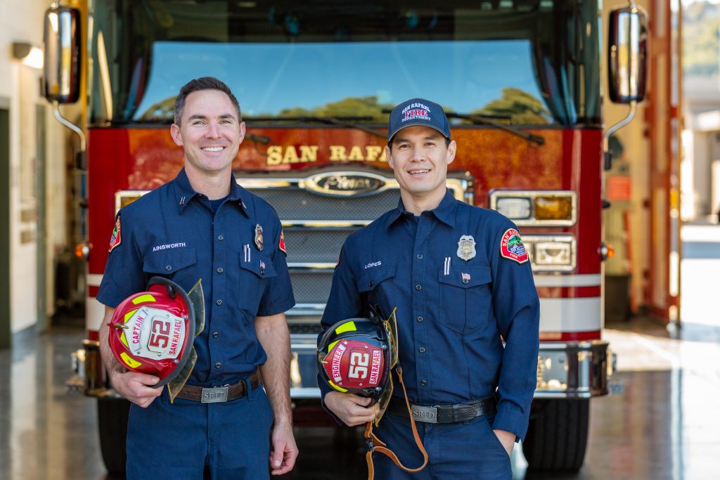 Two smiling San Rafael Fire Department firefighters holding their helmets, in front of a fire engine.