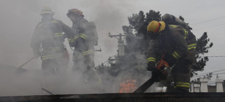 Firefighters on a charred roof. One has a chainsaw that he is using the open the roof.