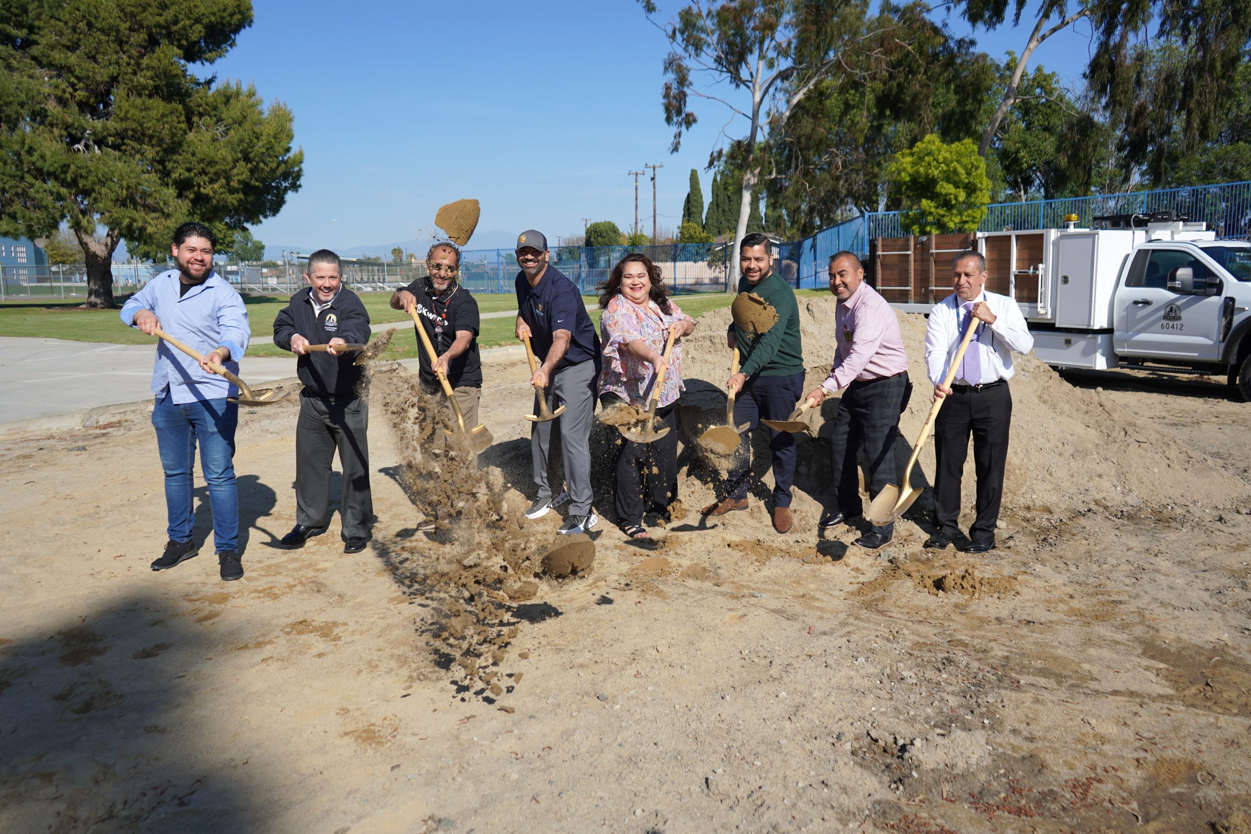 A group of people use shovels to dig and throw dirt into the air on a dirt lot with trees and a white truck behind them.