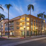 A four-story apartment building on a street corner lined with palm trees and lit with street lights at dusk.