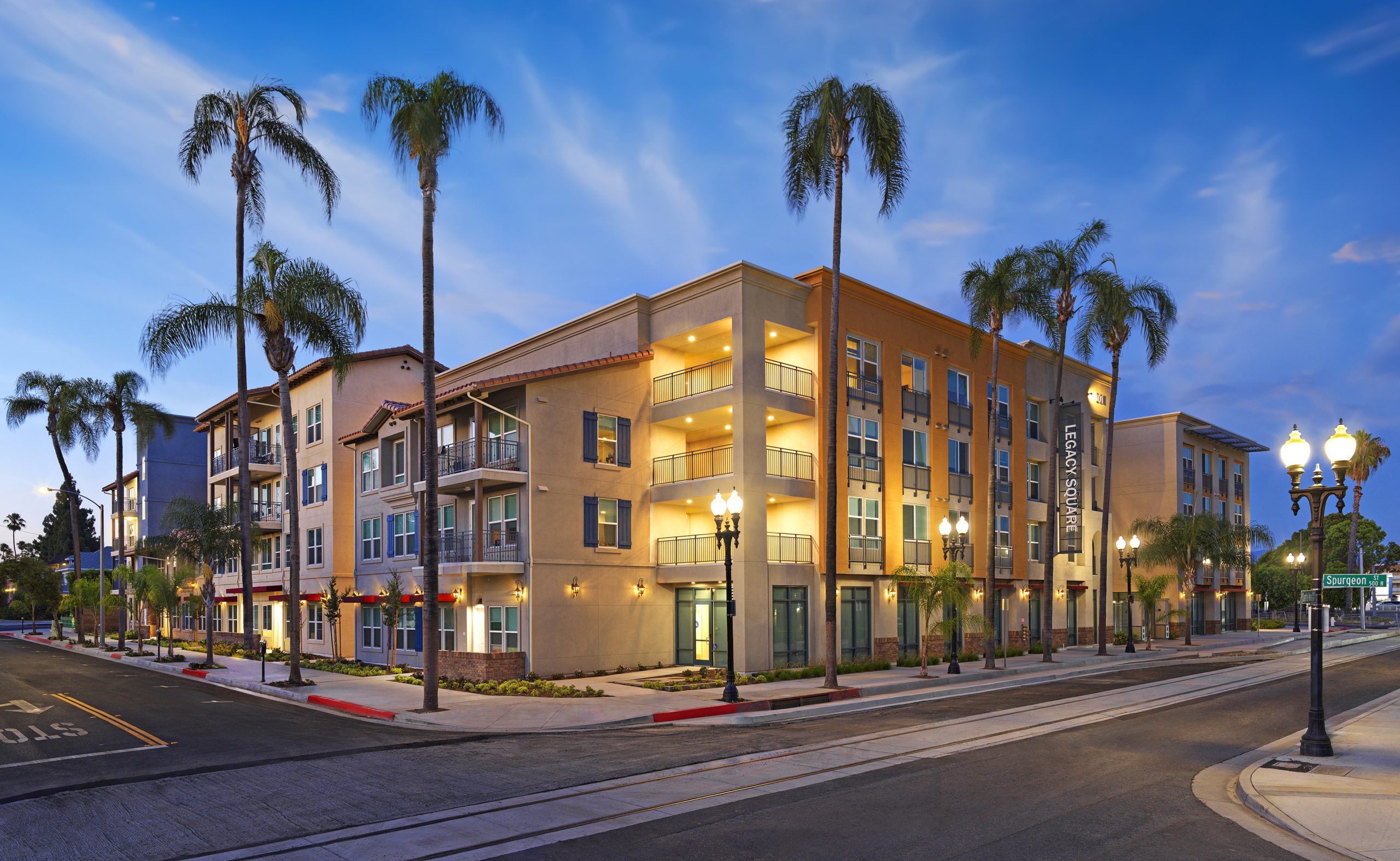 A four-story apartment building on a street corner lined with palm trees and lit with street lights at dusk.