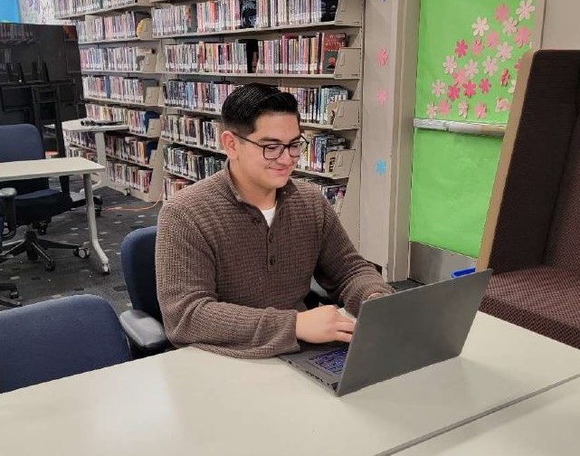 A young man wearing glasses sits at a table using a laptop computer.