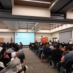 A crowd sits in a room listening to a woman speak behind a podium.