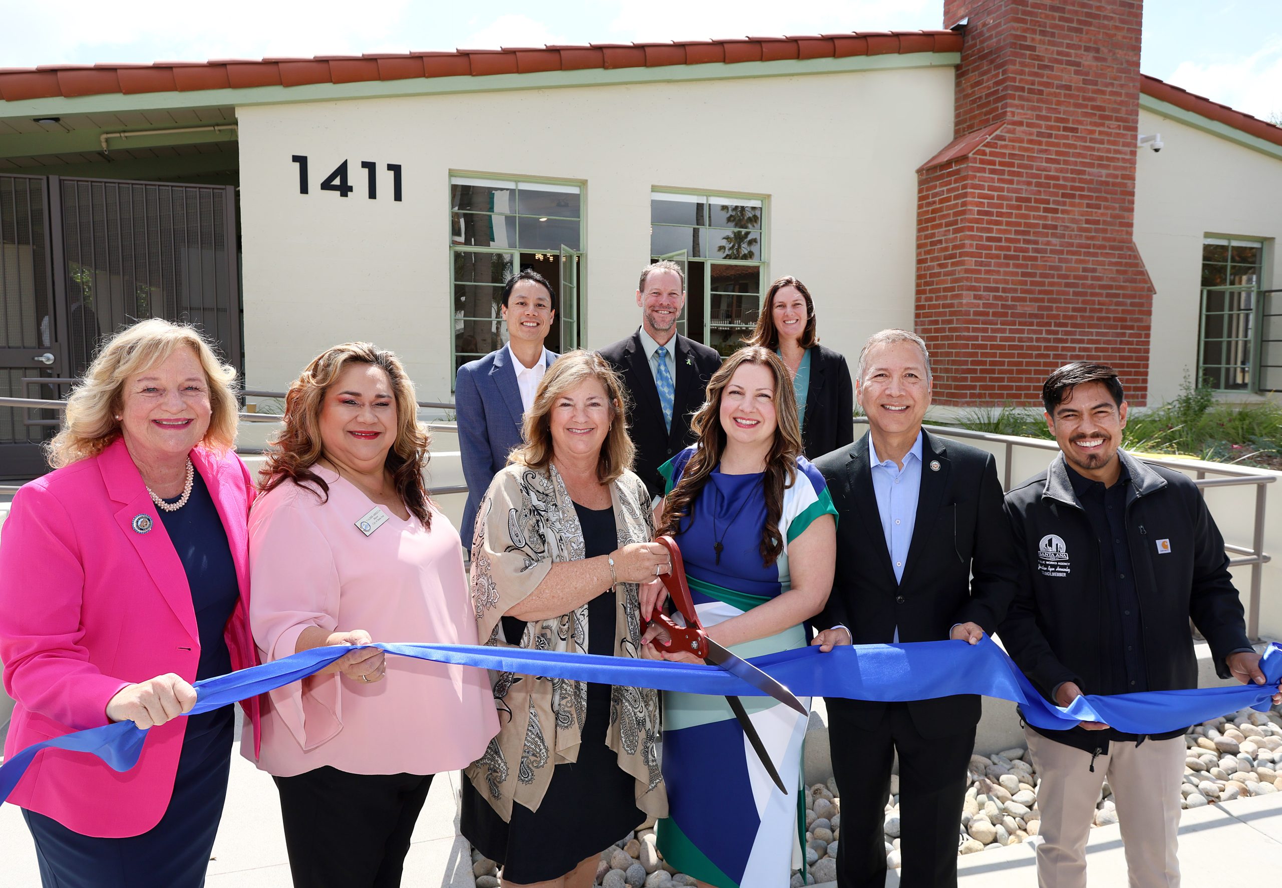 A group of people hold a ribbon with an oversized pair of scissors in front of a building.