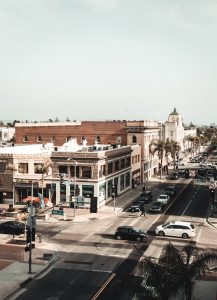 Downtown Santa Ana Rooftop View. A view of Main St and 4th St from the roof of a building, looking south along Main St.. There are cars in the intersection, and two are in the intersection. All of the cars are either black or white. The picture highlights the buildings along Main, including the old City Hall with it's grand marble sides and green roof.