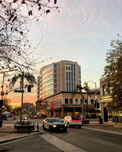 Federal Courthouse. A view of 4th and Broadway at sunset with the Federal Courthouse standing tall and reflecting the warm colors of the sunset. The photo appears to be taken in early fall, as one sycamore tree has lost all it's leaves, but another across the street, outside of Teresa's Jewelers, is still mostly green and only starting to turn brown. In the street there is a police car driving towards the camera and a white crossover SUV waiting at the red light.