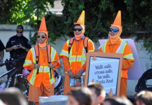 A photograph of the Cone Head Team! Three people stand dressed head to toe in bright orange. They are all wearing safety reflective vests, sunglasses, and cones on their heads as hats. The one on the far right is wearing a sign that reads, "Walk Safe, Cross Safe, Be Safe, Santa Ana!" There are heads of children out of focus in the foreground and there is a police officer in the background.
