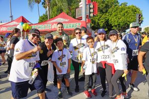 A group of eight runners pose with their Santa Ana 5K Run 2019 shirts and medals. They are all wearing athletic shoes and bottoms, four of them are wearing hats. They're all smiling and looking directly at the camera.