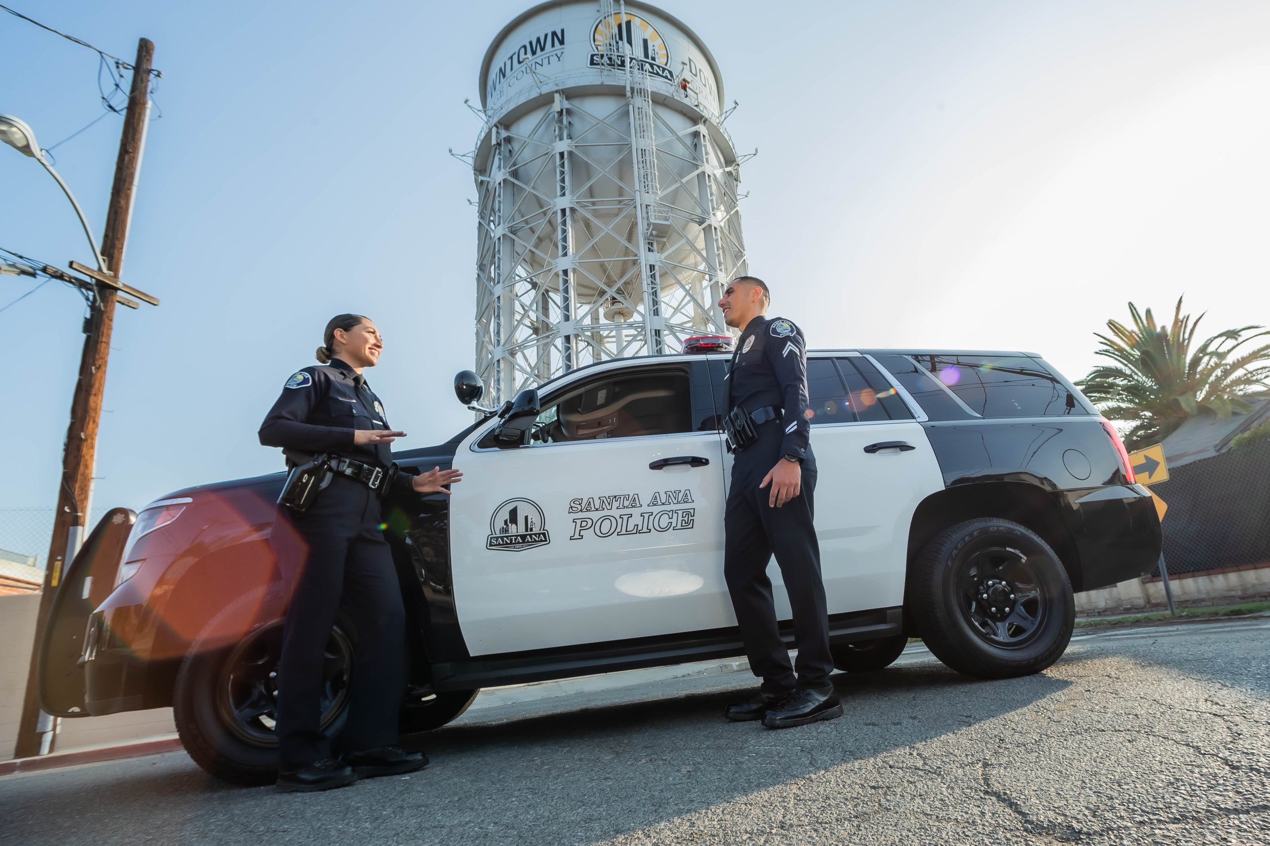 A man and a woman in police uniforms stand in front of a police car under a white water tower.