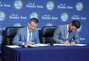 Two men wearing suits and ties sit in black chairs at a blue table in front of a blue background with the City of Santa Ana seal on it and sign papers.