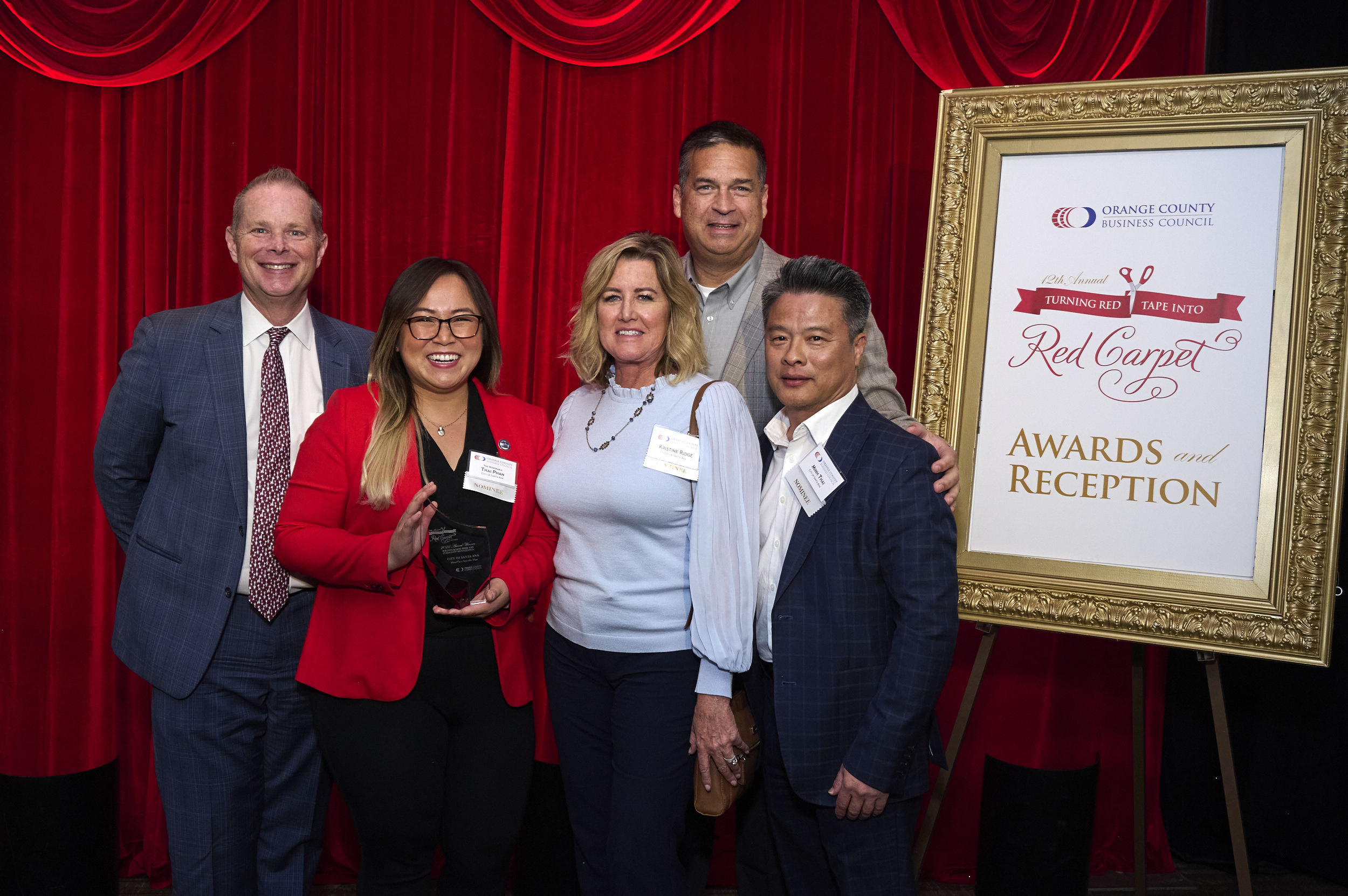 Five people stand in front of a red curtain and a large white sign. A woman in a red coat holds a glass award sculpture.