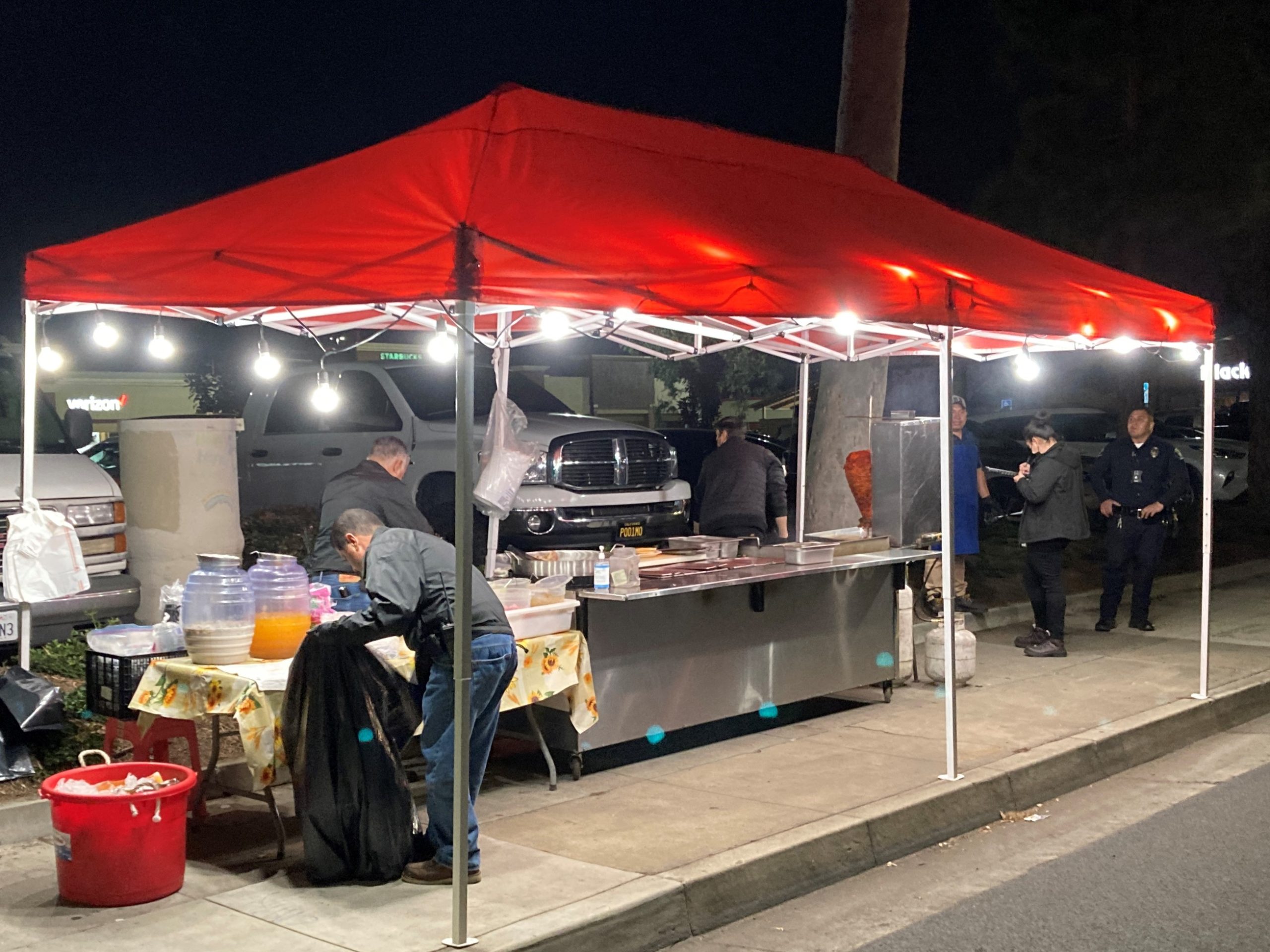 Men remove food from tables under a red tent canopy on a sidewalk at night.