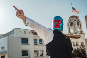 Man wearing a blue and red luchador mask poses in front of the Spurgeon Building