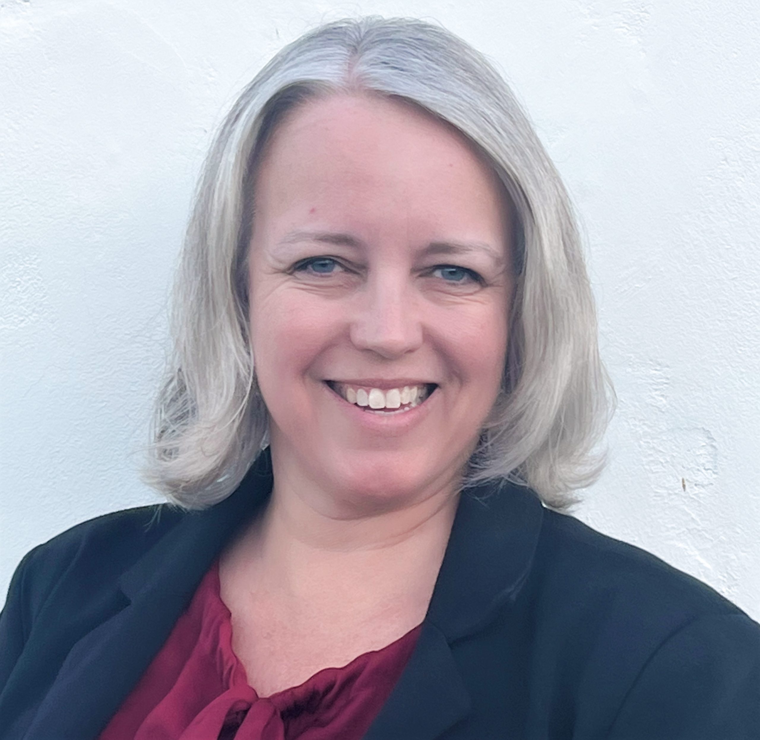 A woman with grey hair wearing a red blouse and blue blazer smiling.