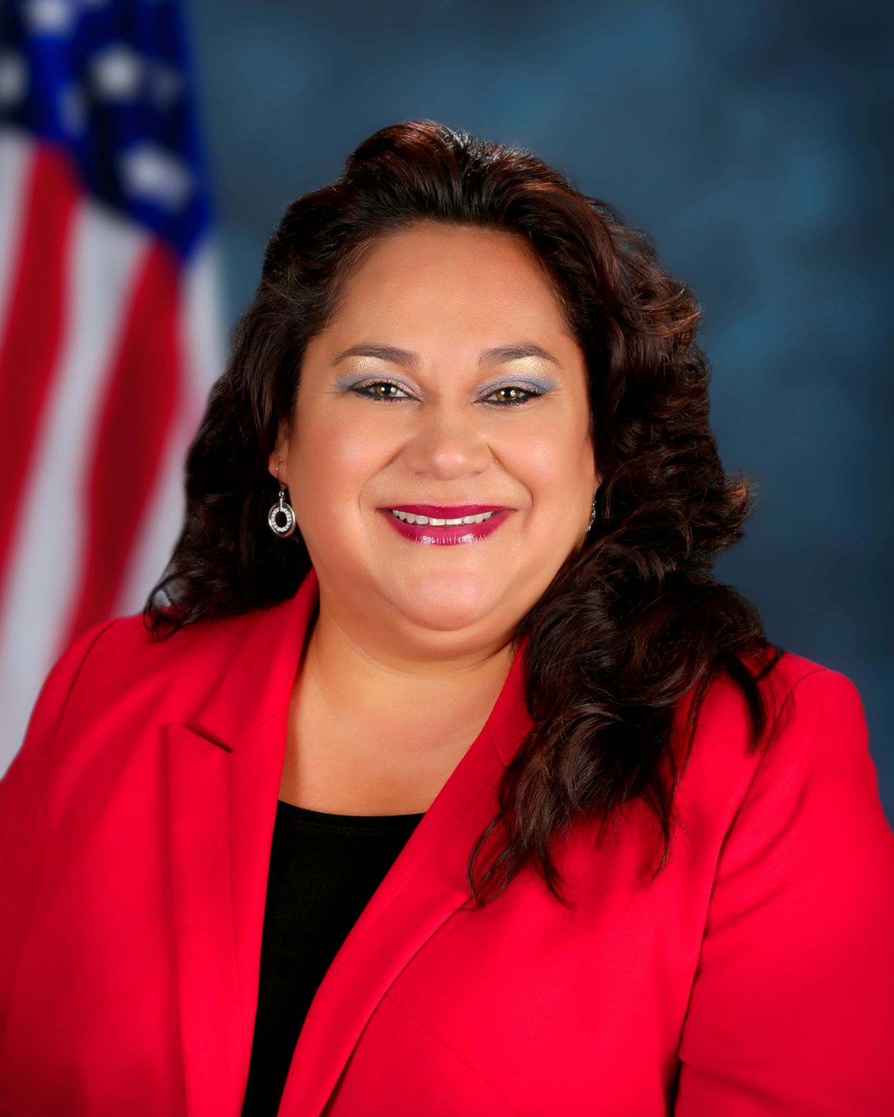 A woman with dark hair wearing a red and black blouse smiles in front of an American flag.