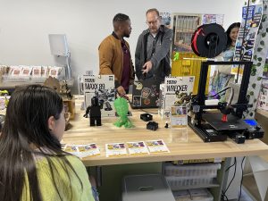 Two men and a girl look at machines and figurines on a wooden table.