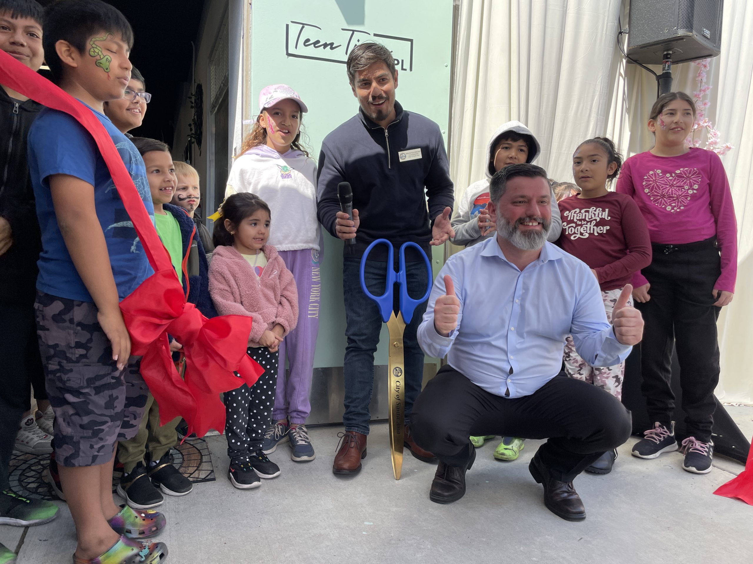 Two men and a group of children smile in front of a sign that says "TeenTime SAPL" next to a large red ribbon that has been cut and a pair of giant scissors.