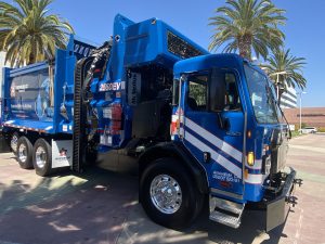 A blue electric trash and recycling collection truck is parking in a roundabout in front of palm trees.