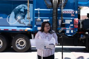 A woman stands behind a clear podium speaking and smiling outside on a sunny day. Behind her is a blue trash collection truck.