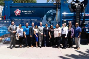 A group of people pose in front of a blue trash collection truck outside on a sunny day.