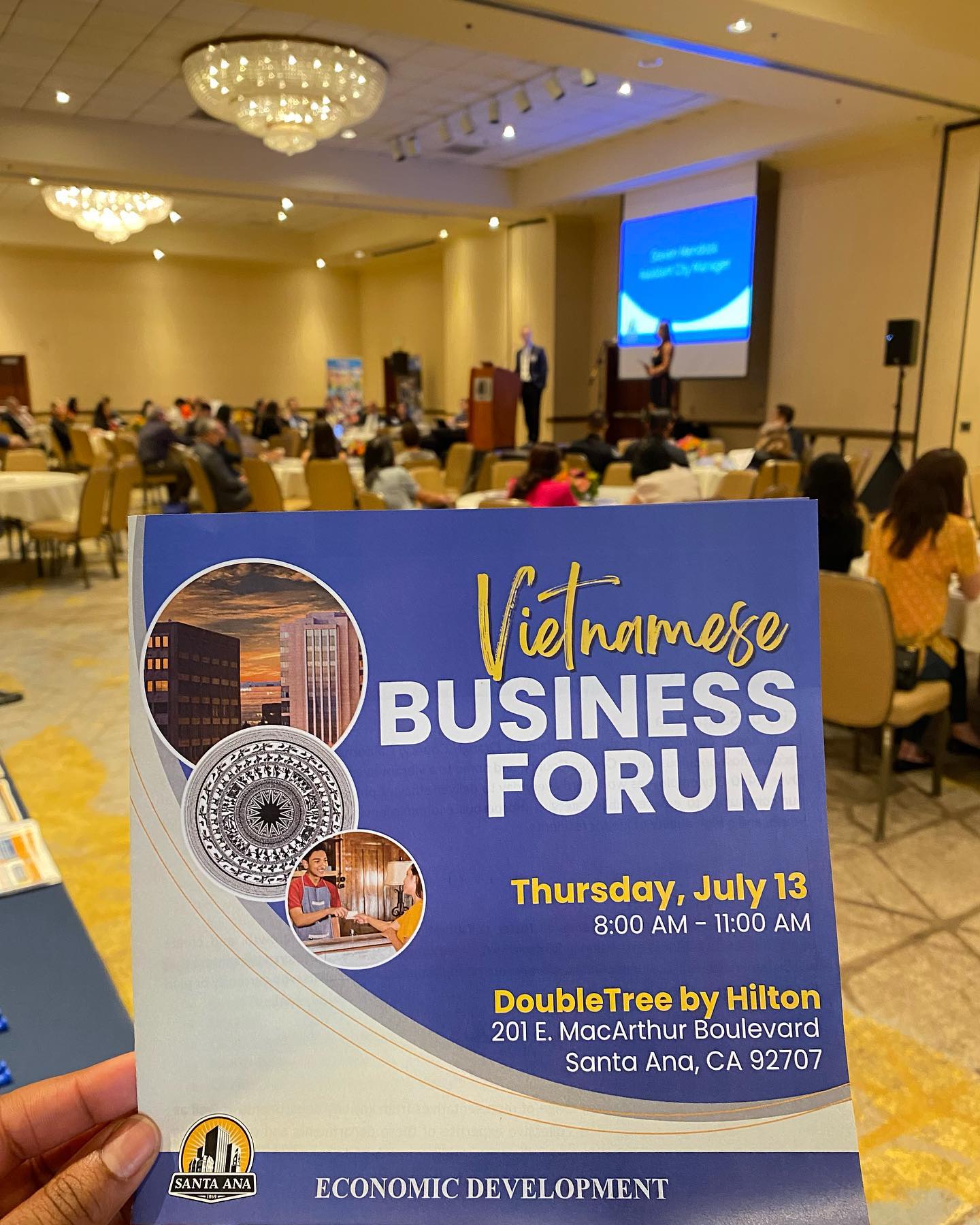 A flyer for the Vietnamese Business Forum is held in front of a crowd of people sitting at tables in a room watching a man speak on a stage in front of a screen.