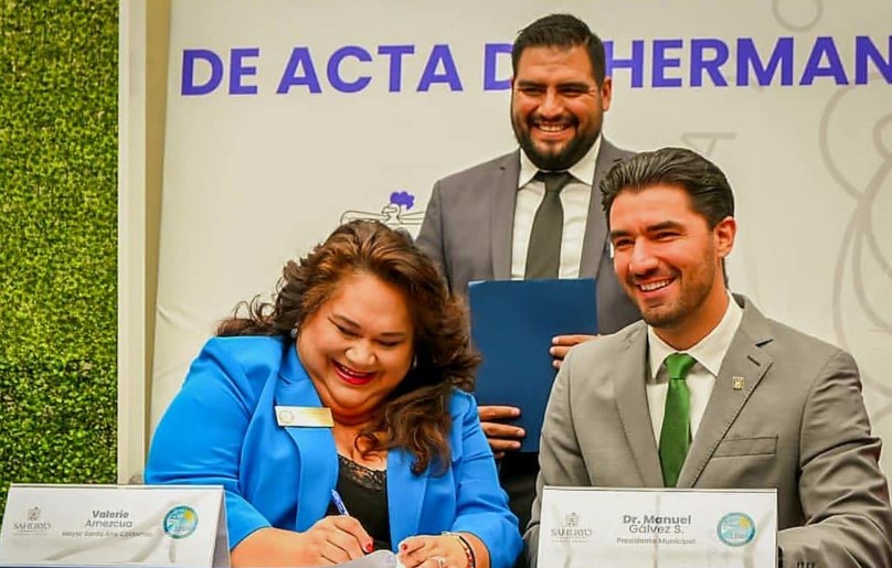 A woman and man sit at a table smiling while the woman signs a piece of paper and another man stands behind them.