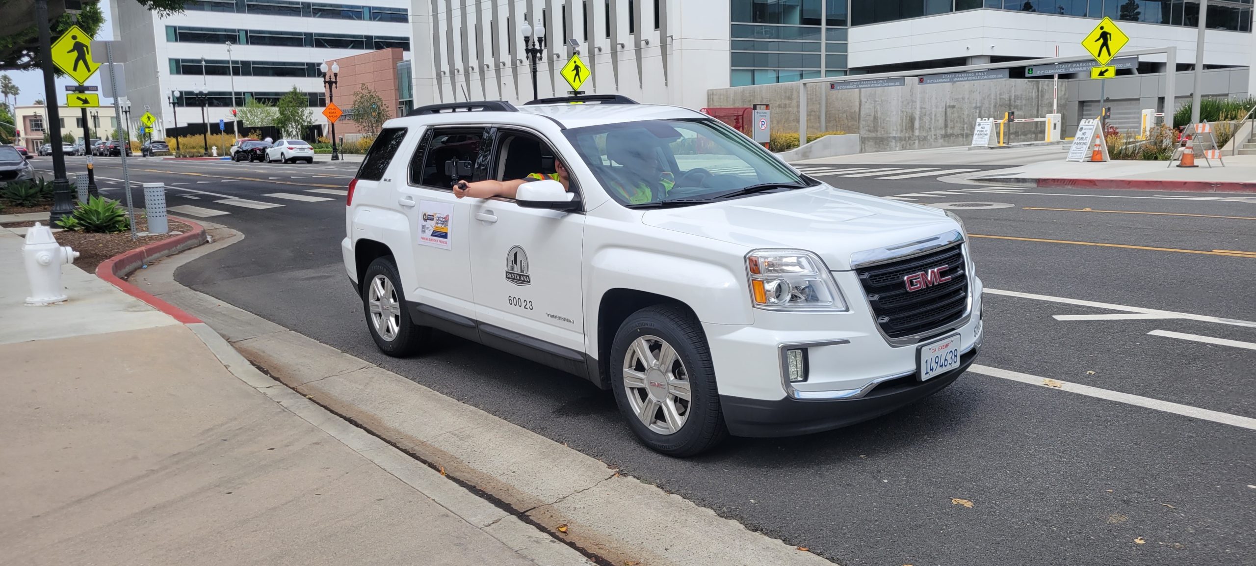 A white vehicle drives on a street while the passenger holds a cell phone out the window.