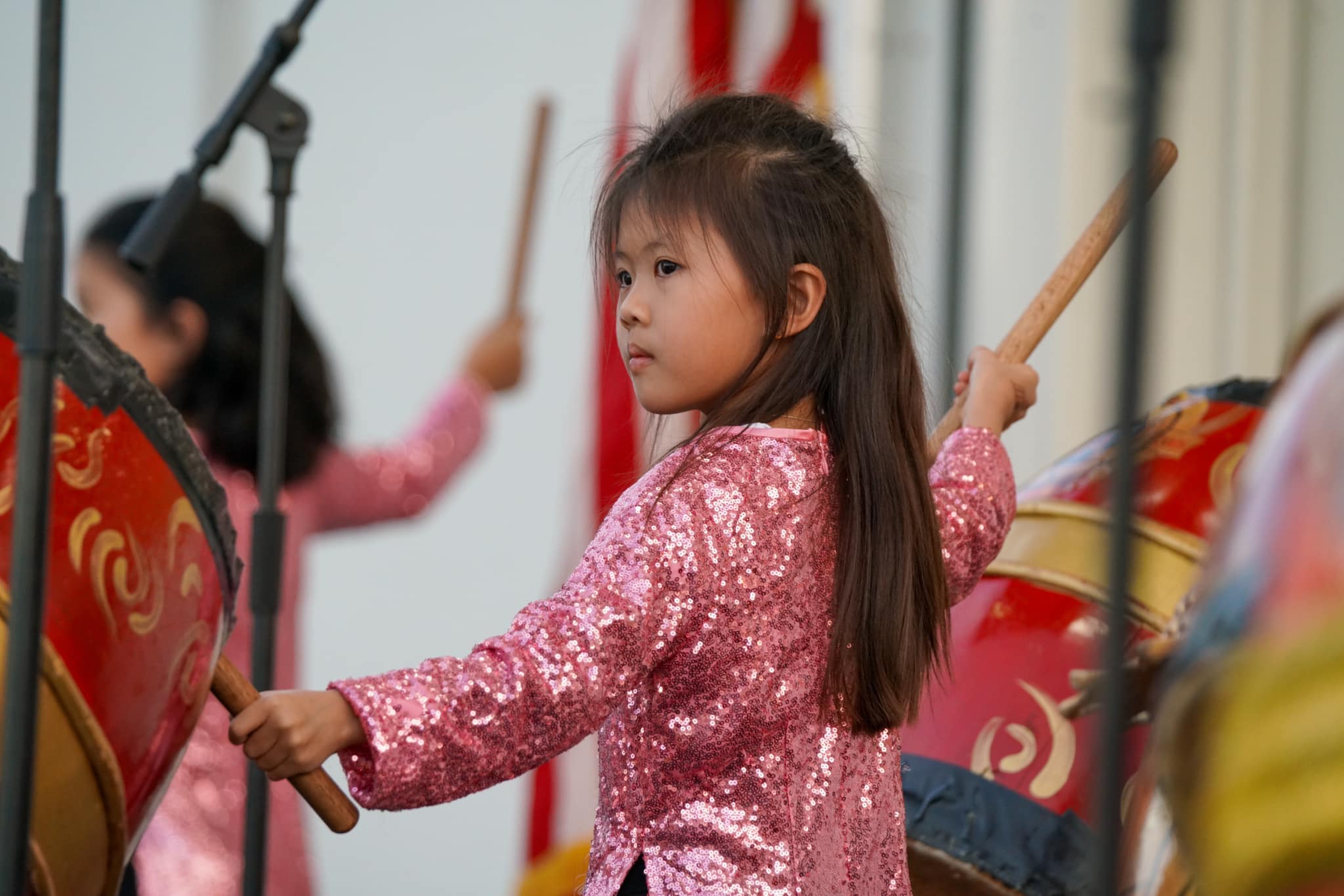 A girl with black hair wearing pink clothing holds a stick while playing a large drum.