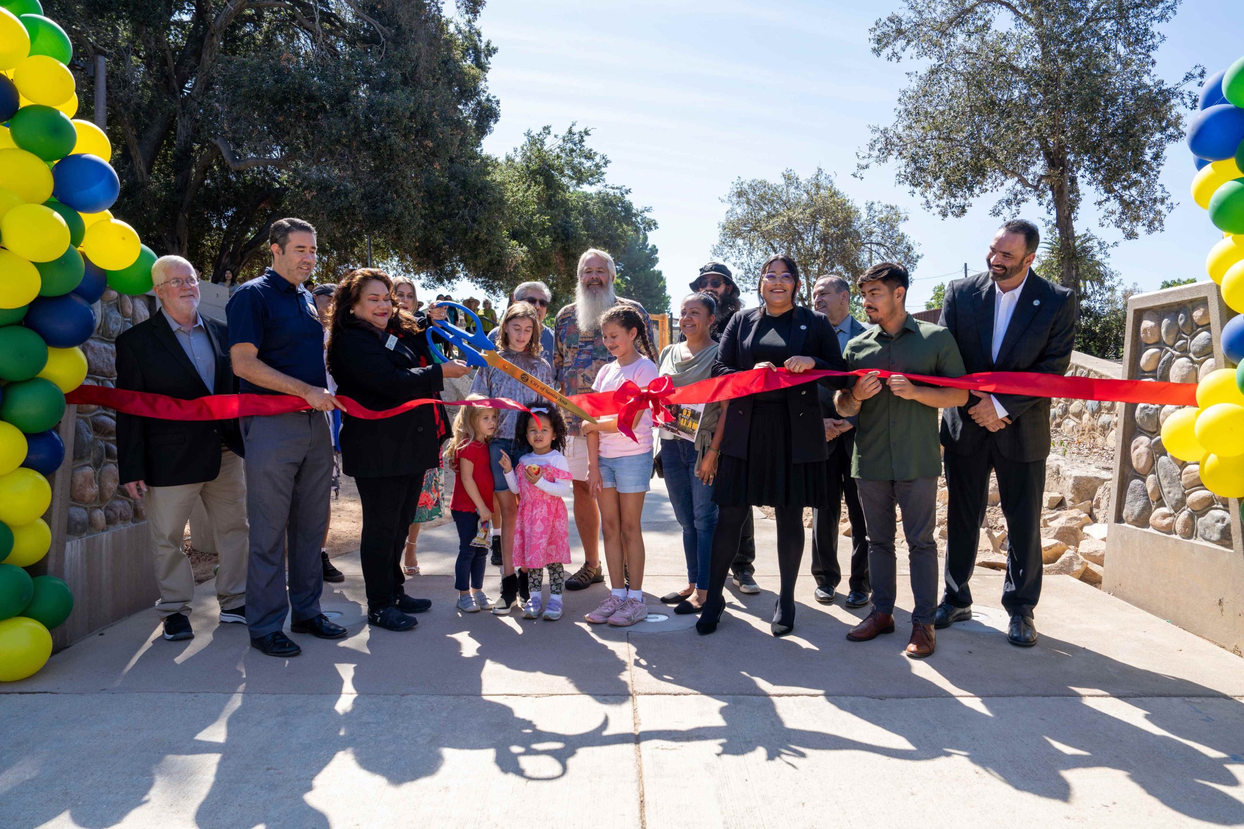 A group of people stand outside in a park with trees, holding a giant red ribbon, while a woman uses large scissors to cut the ribbon.