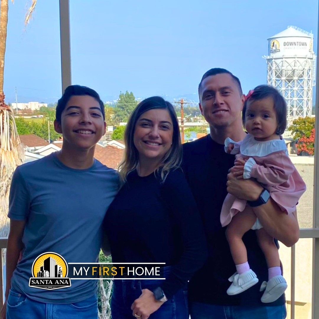 A teenage boy, a woman, and a man holding a young girl stand outside on a porch with a water tower in the background.