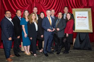 A group of men and women stand together posing and smiling while one man holds an award.