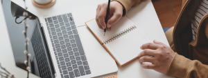 person at desk in front of mac laptop writing on notebook