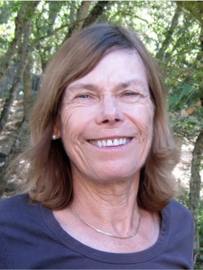 Headshot of Joanne Benjamin, a light-skinned woman with should-length, light brown/blonde hair who is smiling. She is wearing a silver chain necklace and dark blue top. There are trees in the background.