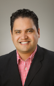 Headshot of Zane Barnes, a man with a tan complexion and black hair who is smiling. He is wearing a red plaid shirt and black suit jacket.