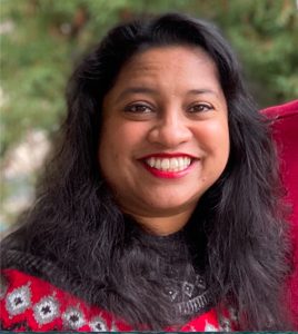 Headshot of Shali Sirkay wearing what appears to be a red winter sweater and matching red lipstick. She has wavy hair that falls just below her shoulders and is standing in front of some kind of foliage.