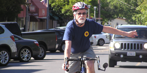 A man wearing a bicycle helmet, riding a bicycle on a street with his hand signaling a left turn with a car behind him.