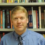 A man in a tie standing in front of a bookcase.