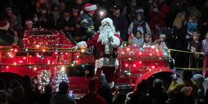 Santa Claus standing on an antique fire truck covered in colored lights.