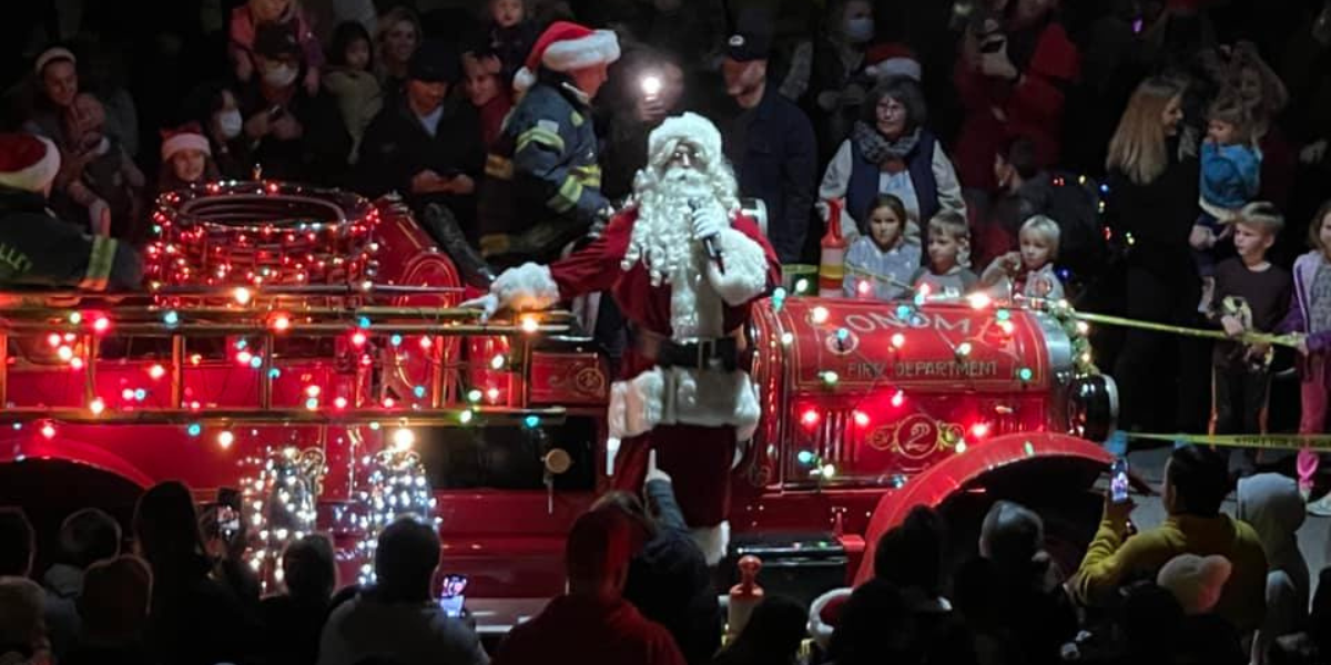 Santa Claus standing on an antique fire truck covered in colored lights.