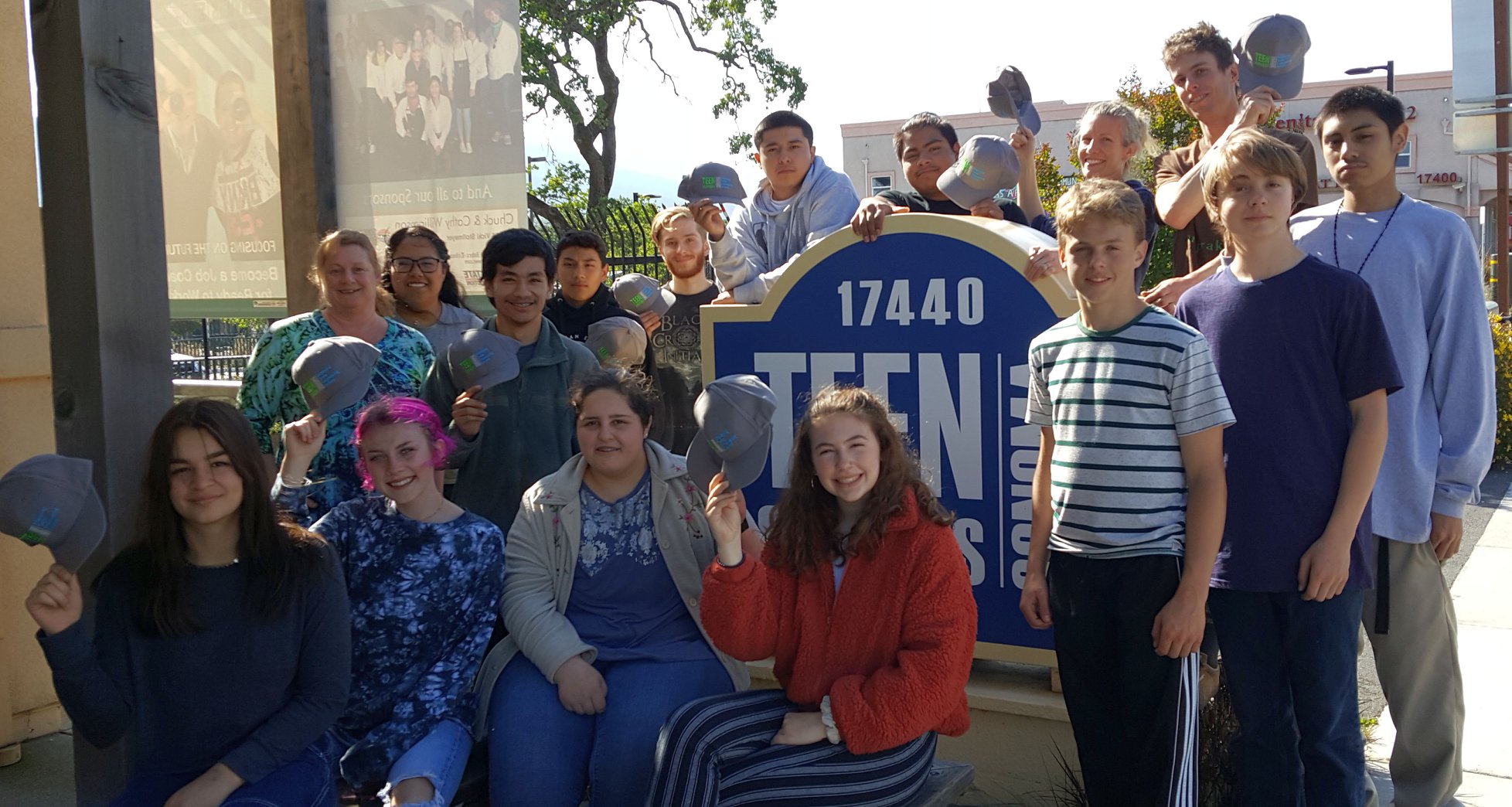 Group of teenagers gathered around the "Teen Services" sign holding up hats with the same logo.