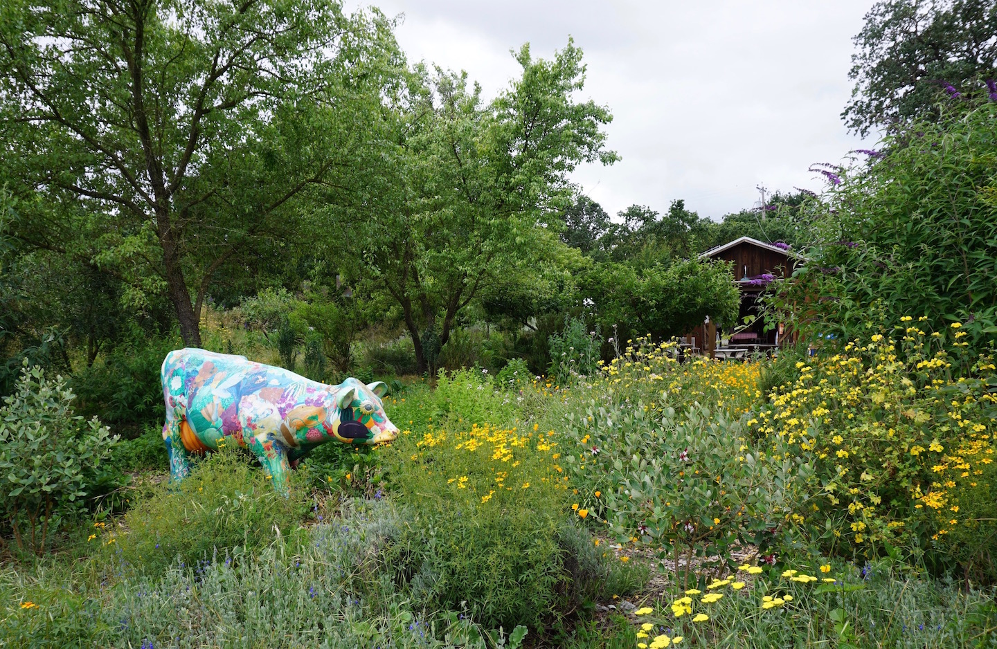 Sclupture of a cow in a garden with flowers and trees with small barn in the background.