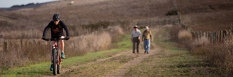 Man riding bike on trail through a field with people walking behind.