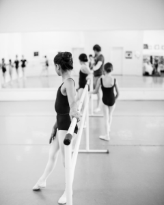 Black and white photo of young ballet dancers warming up on a barre in a dance studio.
