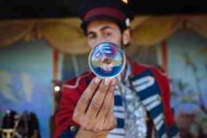 Man in circus barker outfit holding a glass ball with an inverted reflection of himself.