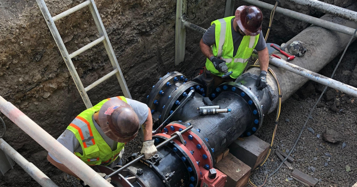 Two workers in safety vests working on a pipeline in a hole.