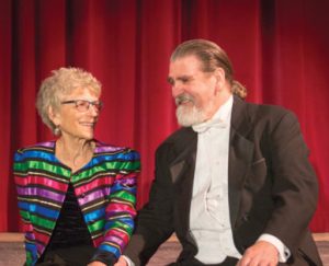 Photo of a woman and a man looking at eachother while sitting on a stage in front of red curtains.