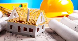 Building plan documents laid out and rolled to the side with a miniature model of a building in the foreground and yellow hard hat in the background.